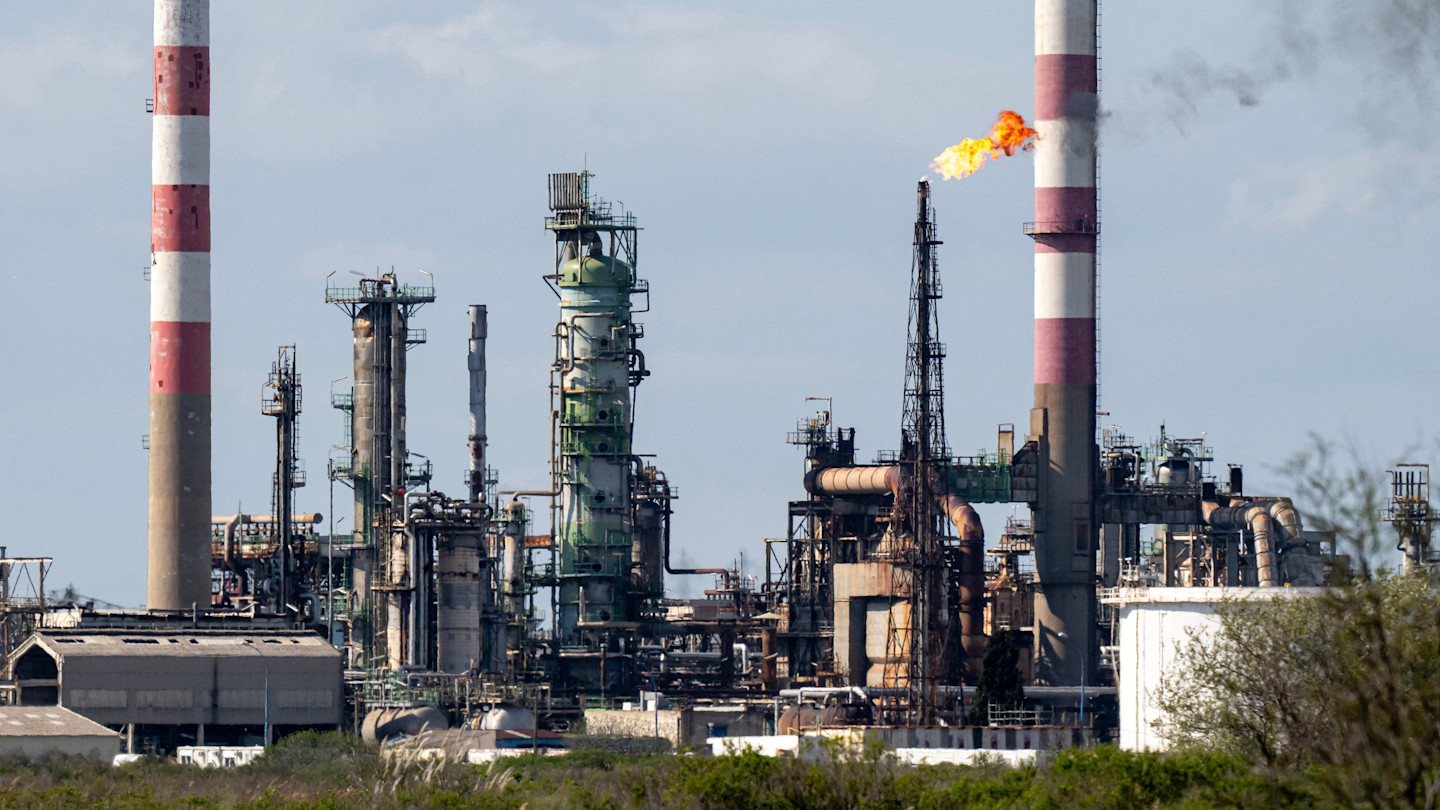 Industrial structures and chimneys at the Esso oil refinery, with a flare burning at the top of one stack.