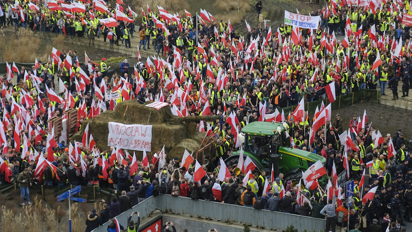 A large crowd of Polish farmers wearing yellow vests waves national flags and gathers around a tractor and hay bales during a protest.