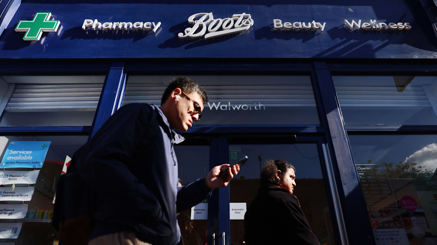 Two pedestrians walk past the entrance of a Boots pharmacy store in London, with the store's signage visible above them.
