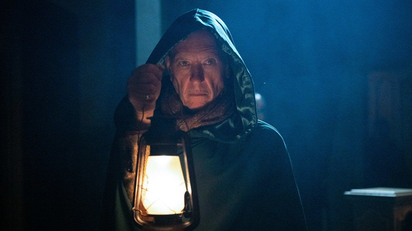 Richard E Grant in a hooded cloak holds a lantern, looking directly ahead in a dimly lit scene from The Celebrity Traitors.