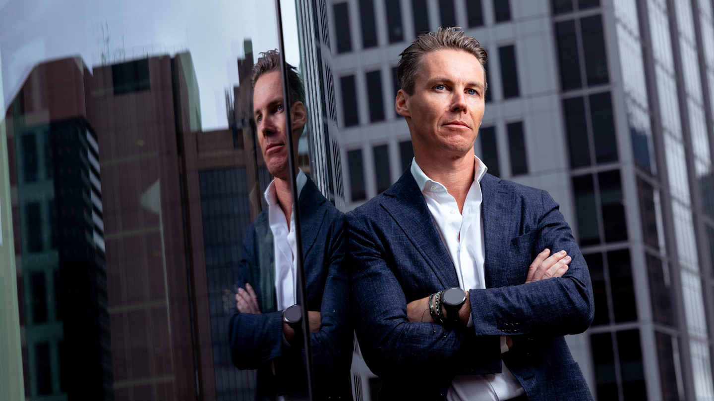 Oliver Curtis stands with arms crossed and is reflected in a glass building facade.