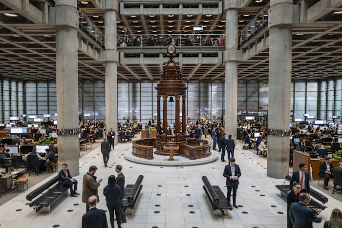 People in business attire work and talk in the main underwriting room at Lloyd’s, with the Lutine Bell at the center.