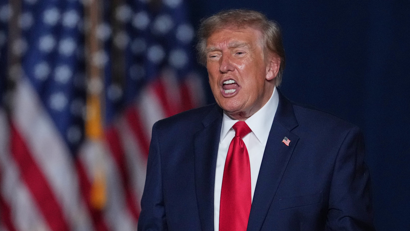 Donald Trump yells with his mouth open at a campaign rally, with American flags visible in the background.