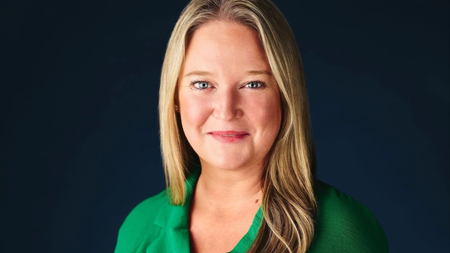 Katie Wiles smiles slightly while wearing a green top against a dark background.