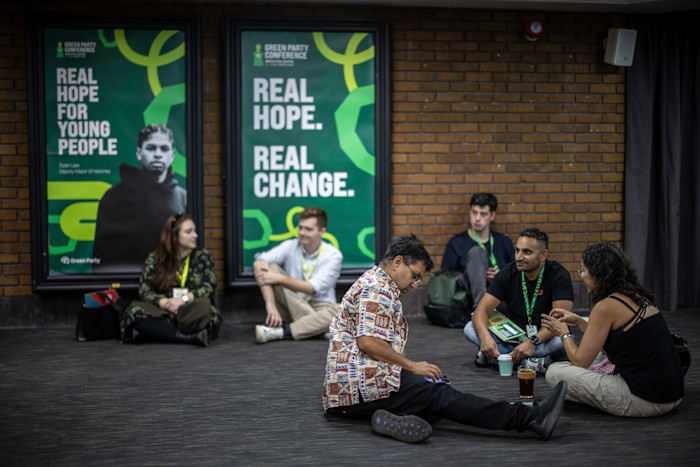 Delegates with lanyards sit on the floor, talking and waiting outside the main auditorium at the Green Party conference, near posters reading “REAL HOPE FOR YOUNG PEOPLE” and “REAL HOPE. REAL CHANGE.”