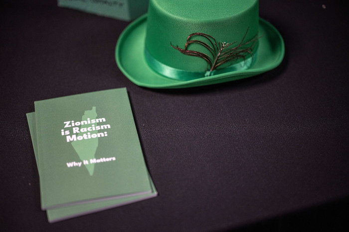 A leaflet titled “Zionism is Racism Motion: Why It Matters” sits on a table next to a green hat with a feather.