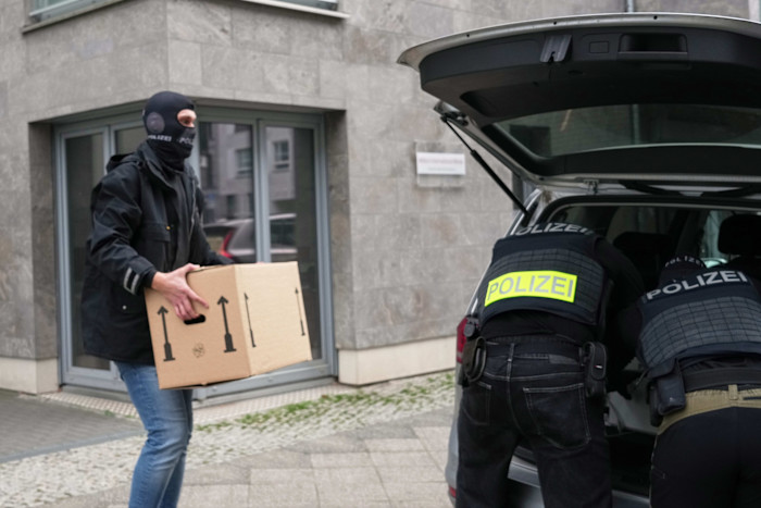 Police officers, some wearing balaclavas and marked vests, load a cardboard box into the back of a vehicle outside a building.