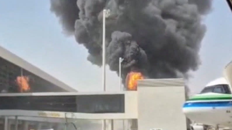 Smoke and flames billow from a building at King Khalid Airport as a plane is parked nearby.