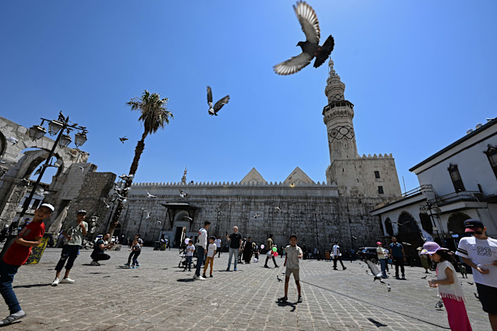 People, including children, walk and feed pigeons in a sunny square near the Umayyad Mosque in Damascus.