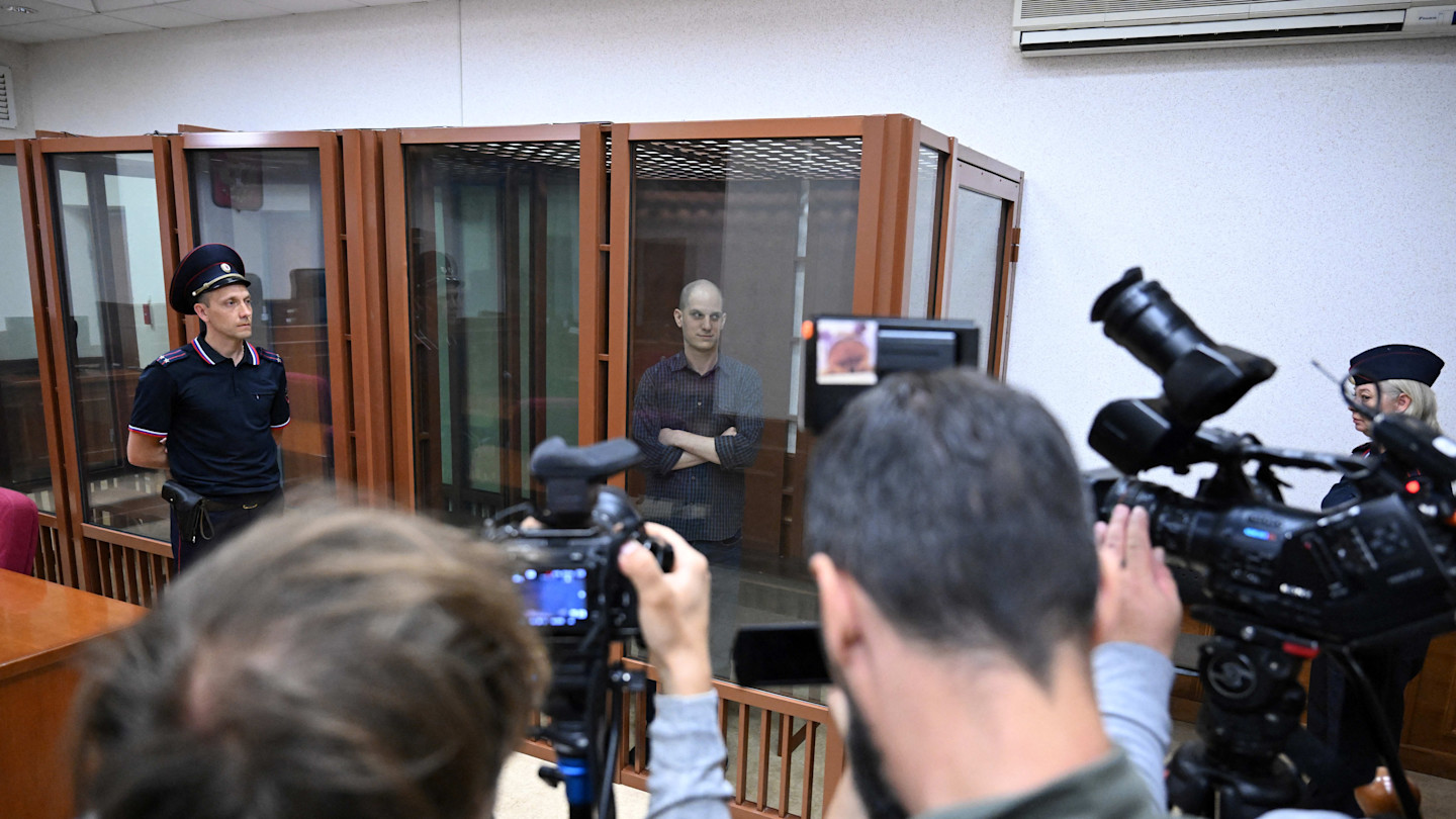 Evan Gershkovich stands inside a glass defendants' cage in court as journalists and security personnel look on.