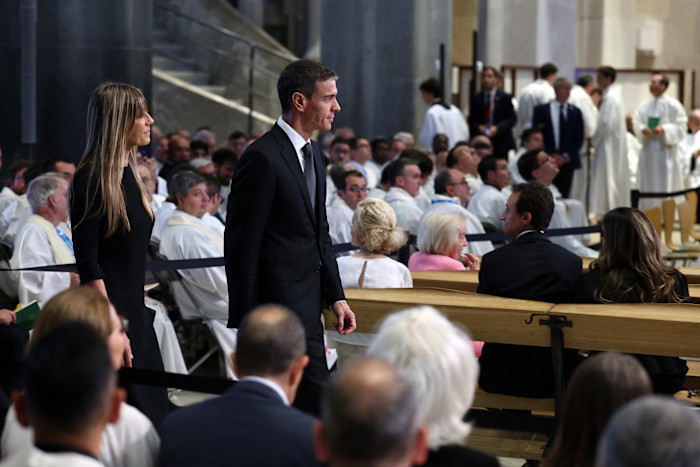 Prime Minister Pedro Sánchez and Begoña Gómez walk down the aisle at a church service, surrounded by seated attendees in white robes.