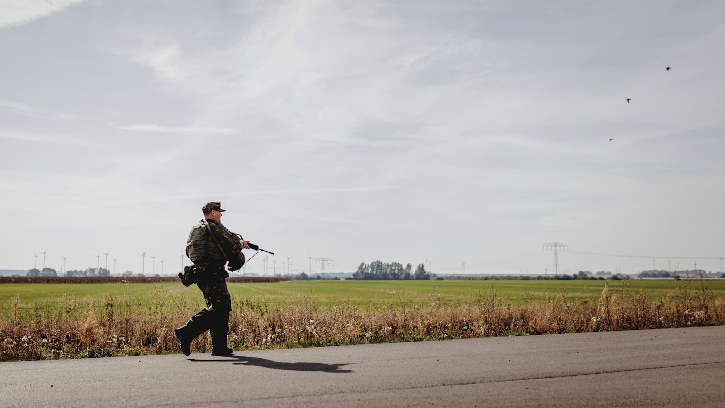 A Bundeswehr soldier in uniform walks along a rural road carrying a rifle during a training exercise.