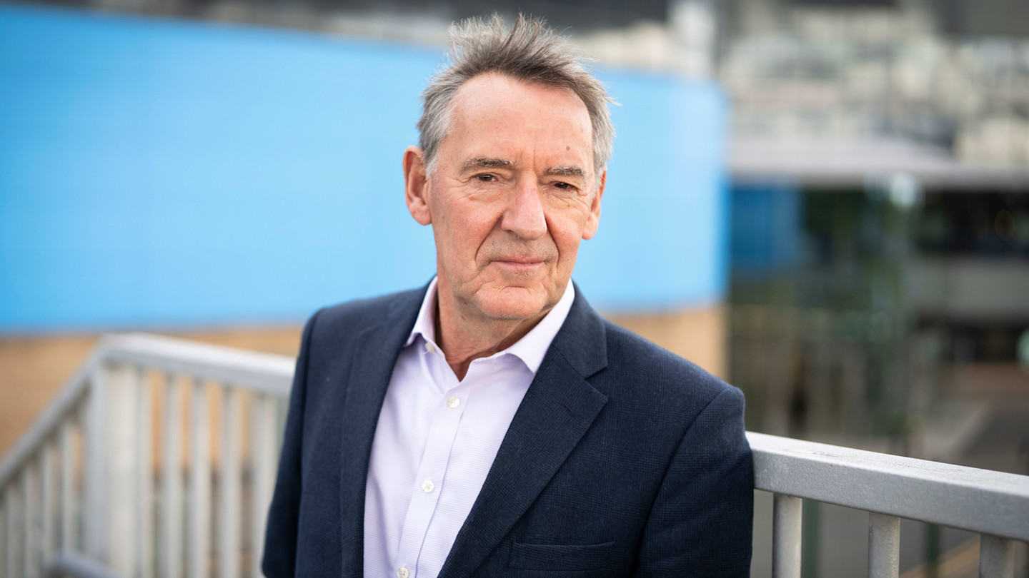 Jim O’Neill stands outdoors in a suit jacket and open-collared shirt, looking directly at the camera.