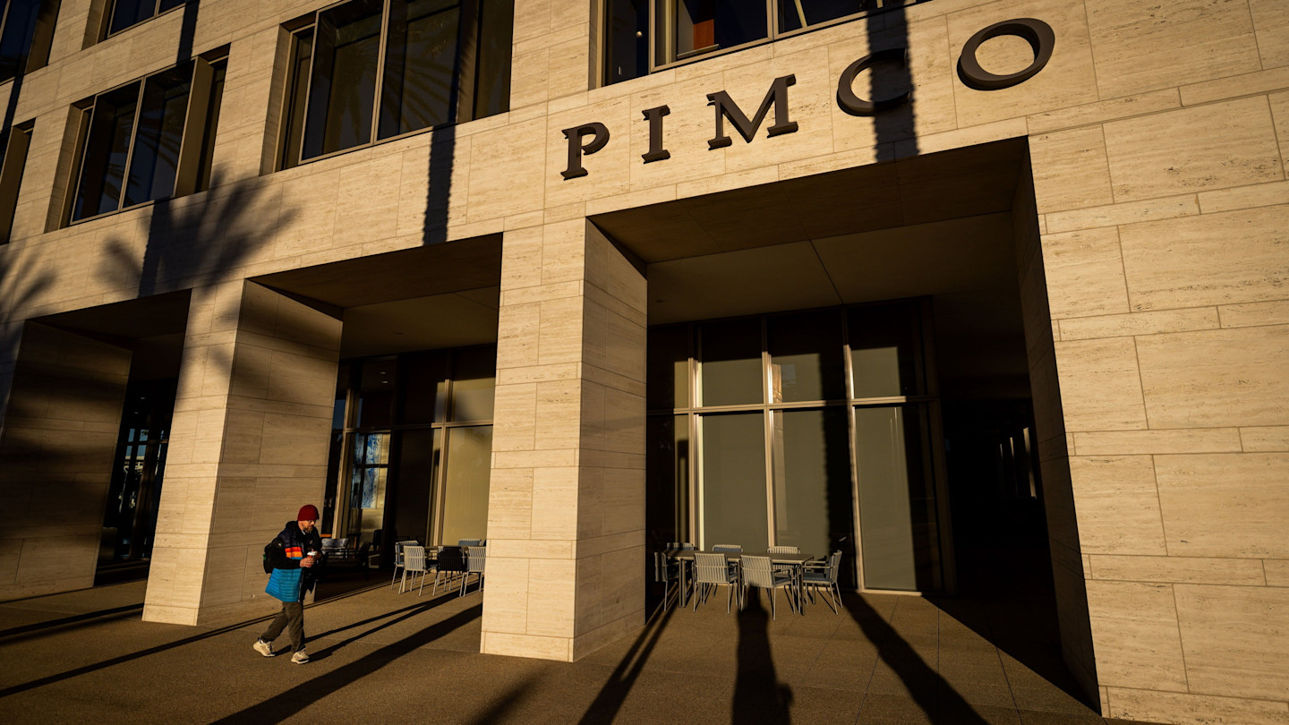 Pimco headquarters building exterior with large windows and pillars, shadowed by nearby palm trees. A person walks past the entrance.