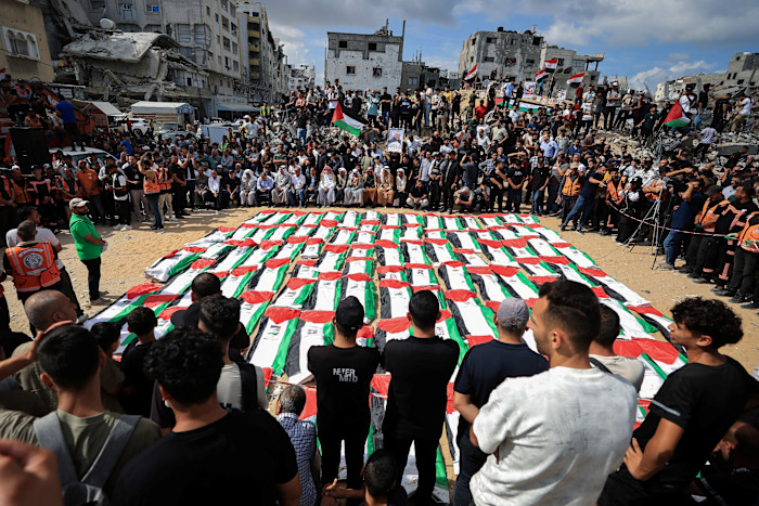 Rows of bodies covered in Palestinian flags are surrounded by mourners at a mass funeral in Gaza City amid damaged buildings.