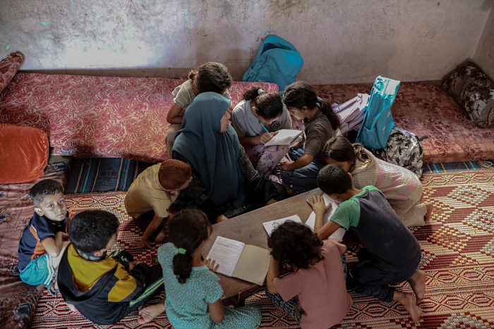 Children sit closely together around a low table inside a shelter, reading and writing in notebooks, with sleeping mats and backpacks nearby.