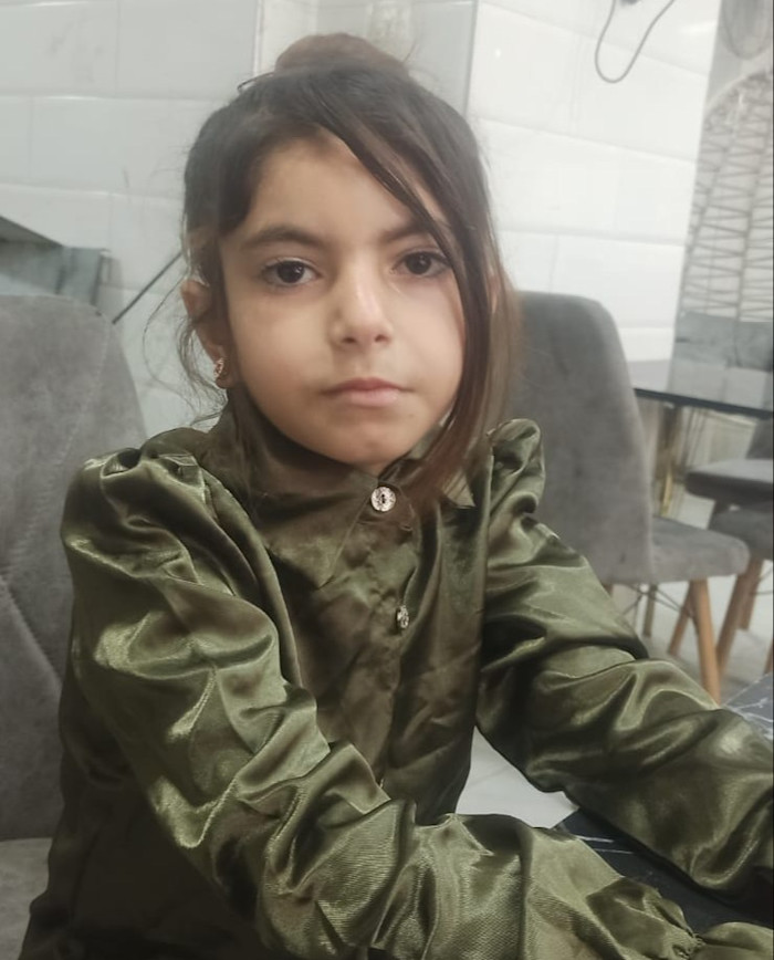 Retaj, a young girl with long brown hair, sits at a table wearing a shiny green blouse and looks directly at the camera.