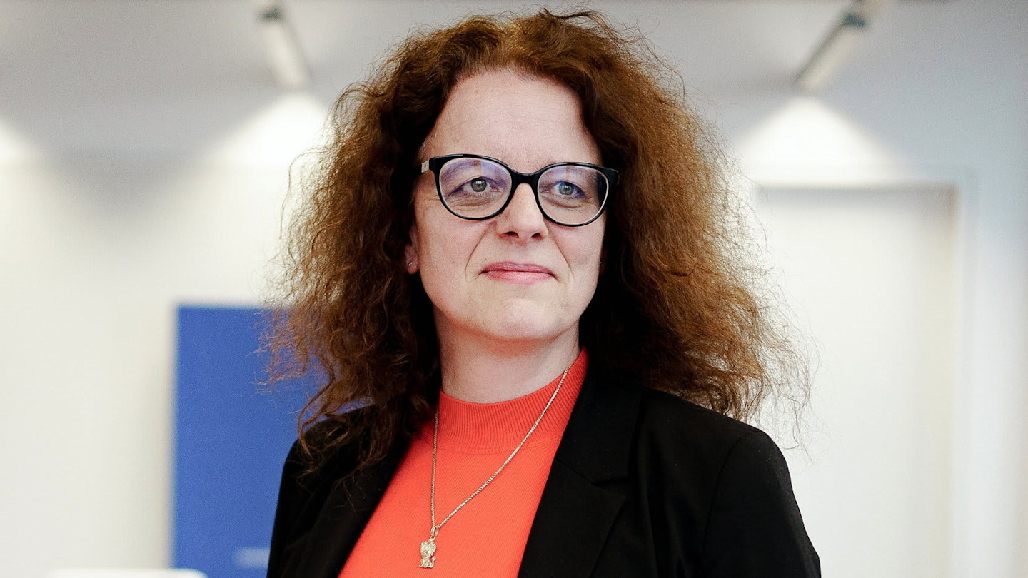Isabel Schnabel stands in an office wearing glasses, a black blazer, and a red top with a pendant necklace.