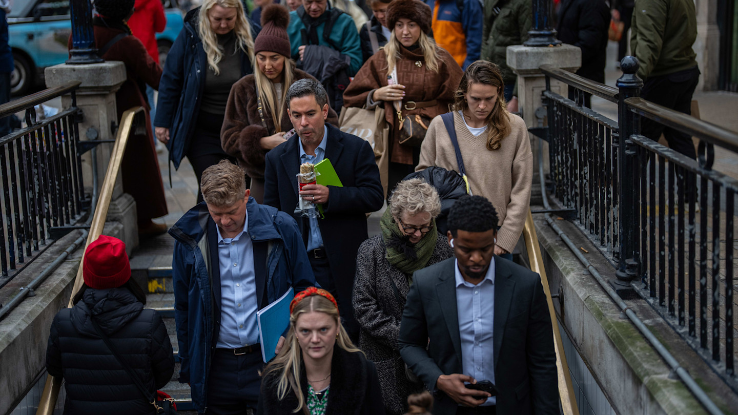 Commuters walk down stairs into Oxford Circus Tube station, some looking at phones or holding drinks and shopping bags.