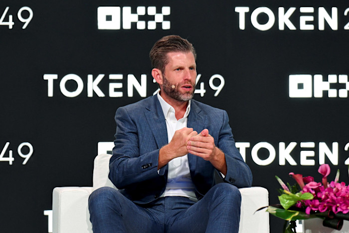 Eric Trump speaks on stage at the TOKEN2049 conference, gesturing with his hands while seated in a chair.