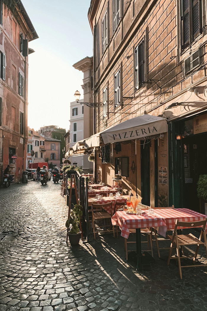 Outdoor tables with red and white checkered tablecloths are set at a pizzeria along a cobblestone street in Trastevere, Rome.