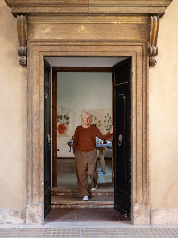 Isabella Ducrot stands in the doorway of her studio, with several of her artworks visible on the wall behind her.
