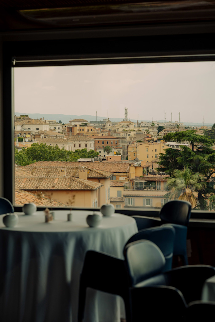 A round table set for dining is positioned by a window with a view of Rome’s rooftops and distant buildings.