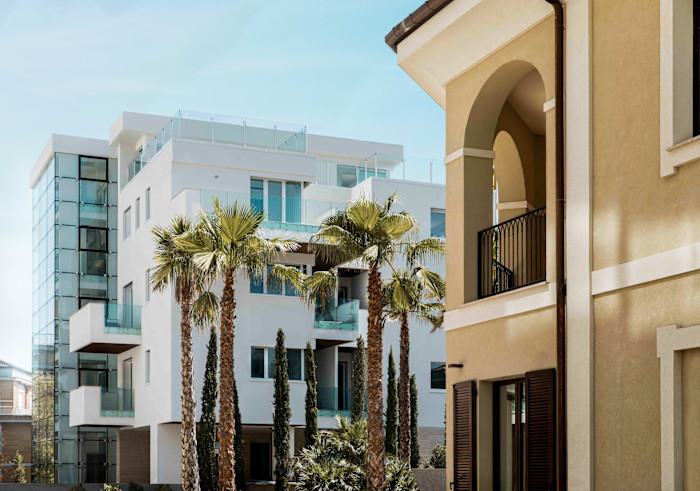 Modern white apartment building with large balconies and glass railings, palm trees in front, and a tan building with arched balconies on the right.