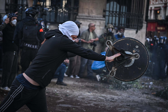 A protester with a covered face pushes a large metal object during a clash at a student demonstration in Paris. Riot police and press are visible in the background.