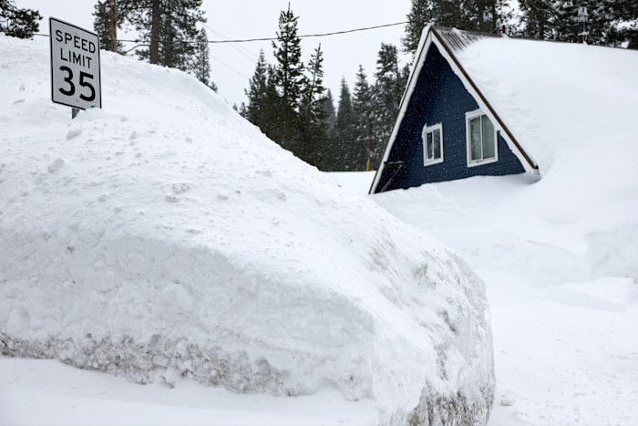 A house and a speed limit sign are partially buried by deep snow after a heavy snowstorm, with snow piled high around them.