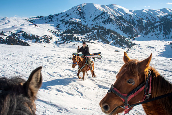A person on horseback carries skis across a snowy mountain landscape in Kyrgyzstan, with two other horses in the foreground.