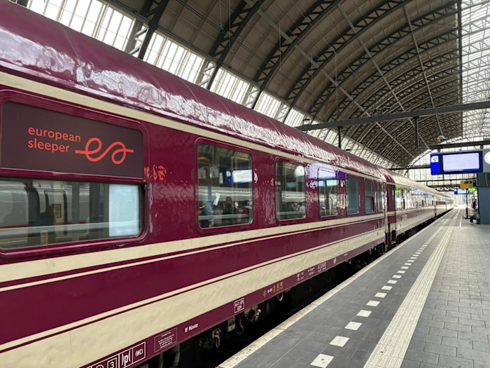 A maroon European Sleeper overnight train is stationed at a covered platform in a large railway terminal.