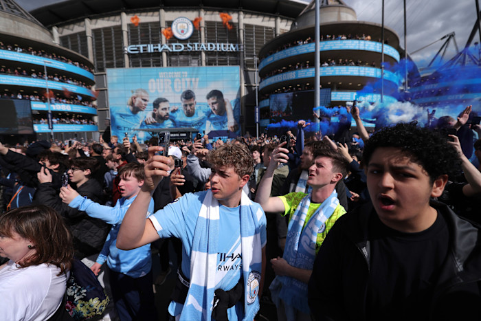 Manchester City supporters gather outside Etihad Stadium, many holding phones and wearing team colors, with blue smoke in the air.