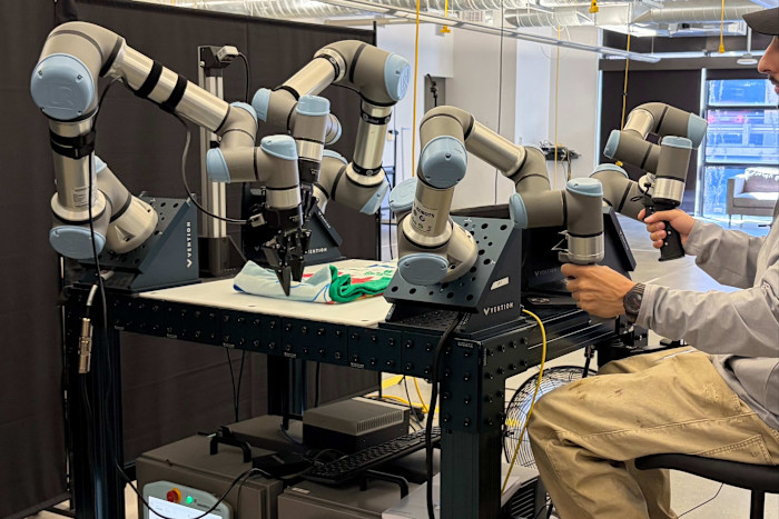 A person operates two handheld controllers to guide robotic arms in folding a piece of clothing on a table.