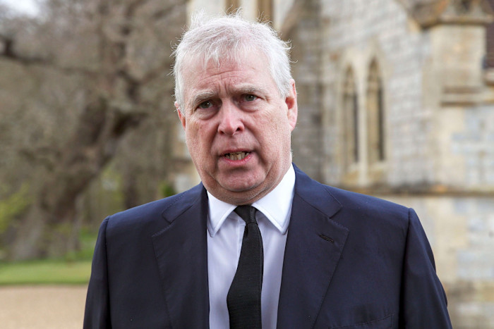 Andrew Mountbatten-Windsor speaks outdoors, wearing a dark suit and tie, with a serious expression.