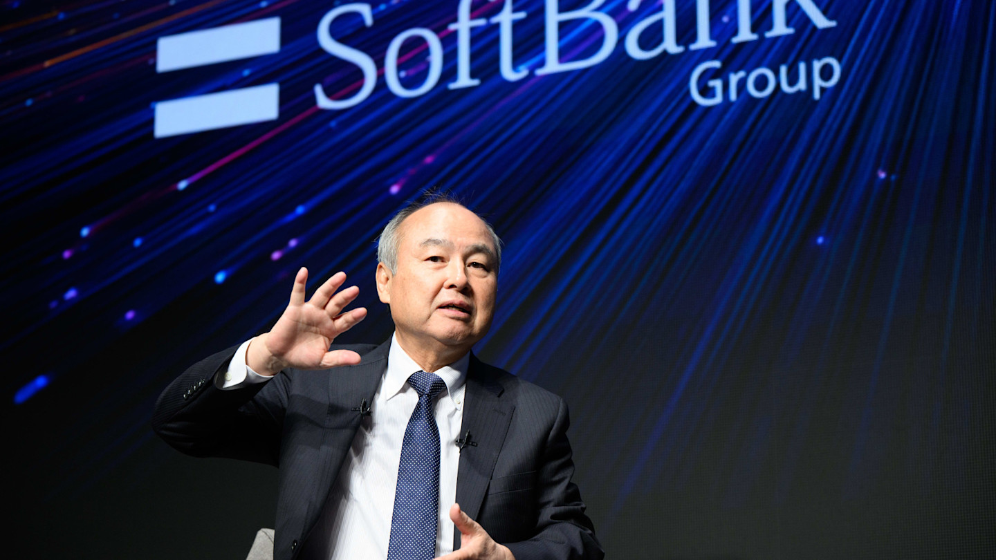 Masayoshi Son gestures while speaking at a news conference, with the SoftBank Group logo displayed behind him.