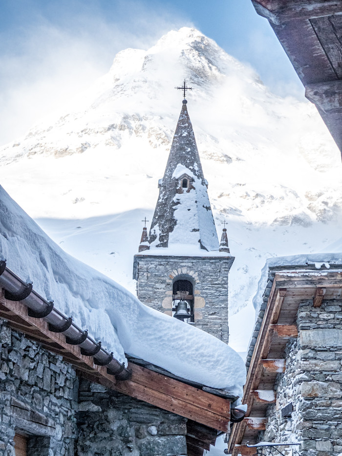 The stone bell tower of the church in Bonneval-sur-Arc A snow-covered stone church tower framed by village rooftops and mountains.
