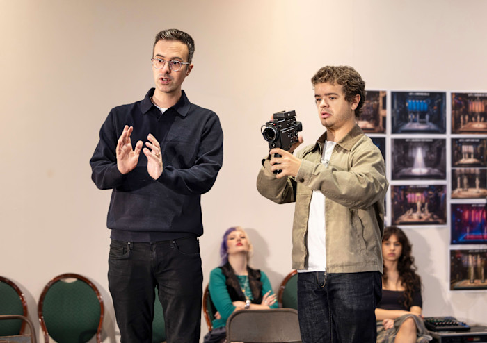 A tall young man in glasses and dark blue top gestures as another young man at his side points a video camera in the same direction.