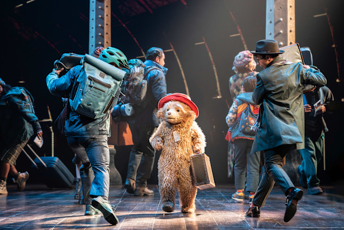 Paddington Bear, wearing a red hat and holding a suitcase, stands on stage surrounded by actors dressed as travelers in a busy train station scene.