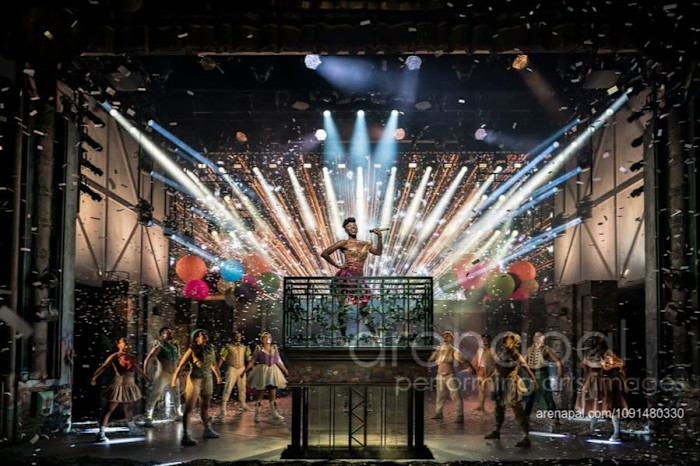 An actress brandishes a microphone, singing on a balcony on stage, surrounded by dancers and dramatic stage lighting with confetti falling.