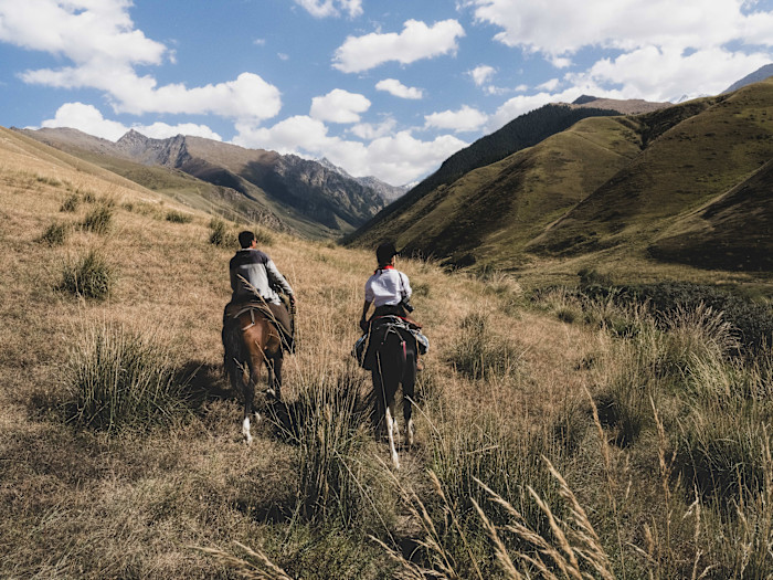 Adil and another person riding horses side by side through grassy hills with mountains in the distance.