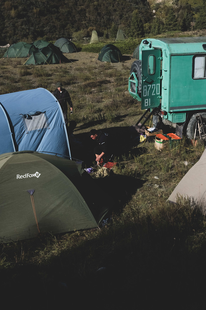 Blue kitchen tent on the left and green truck on the right, surrounded by other camping tents and two people.