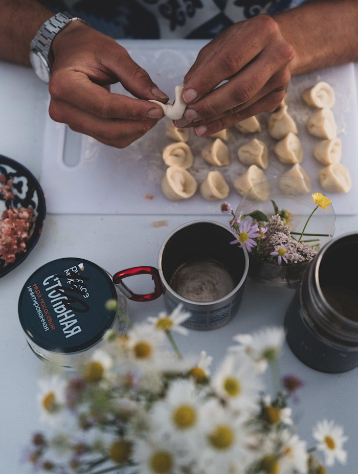 Hands shaping pelmeni dough over a cutting board with several formed dumplings, surrounded by camping mugs, wildflowers, and ingredients.