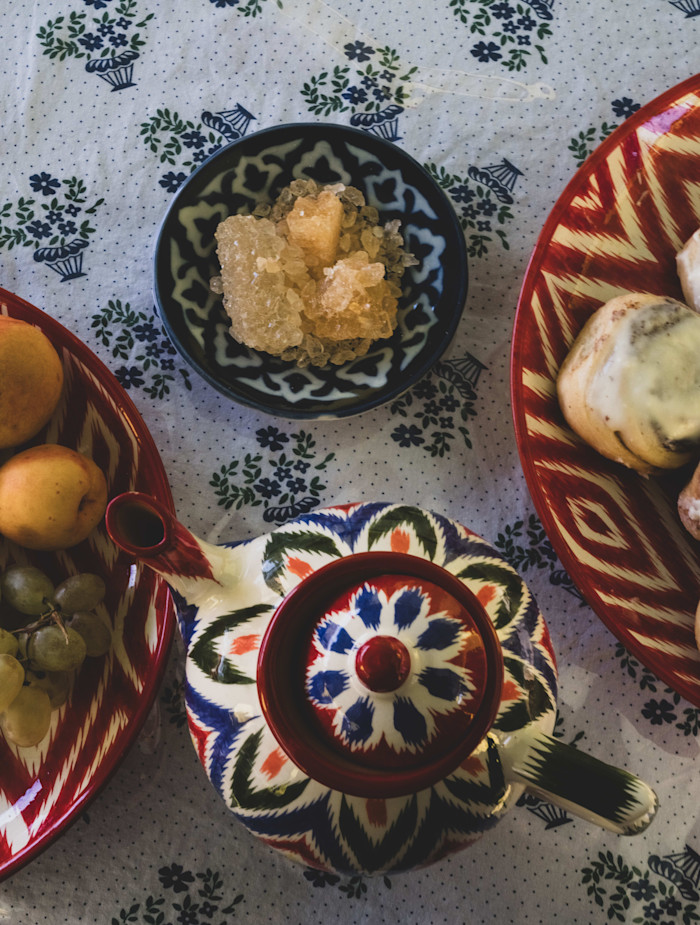 A patterned teapot, a bowl of rock sugar, and colourful plates with cakes and fruit are arranged on a floral tablecloth.