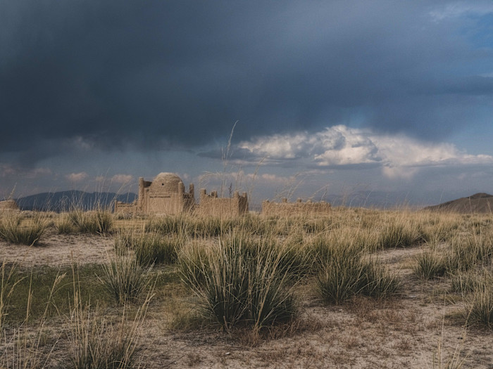 Ancient mausoleums made of earth or stone stand amid dry grass under a dramatic sky with dark clouds and evening light.