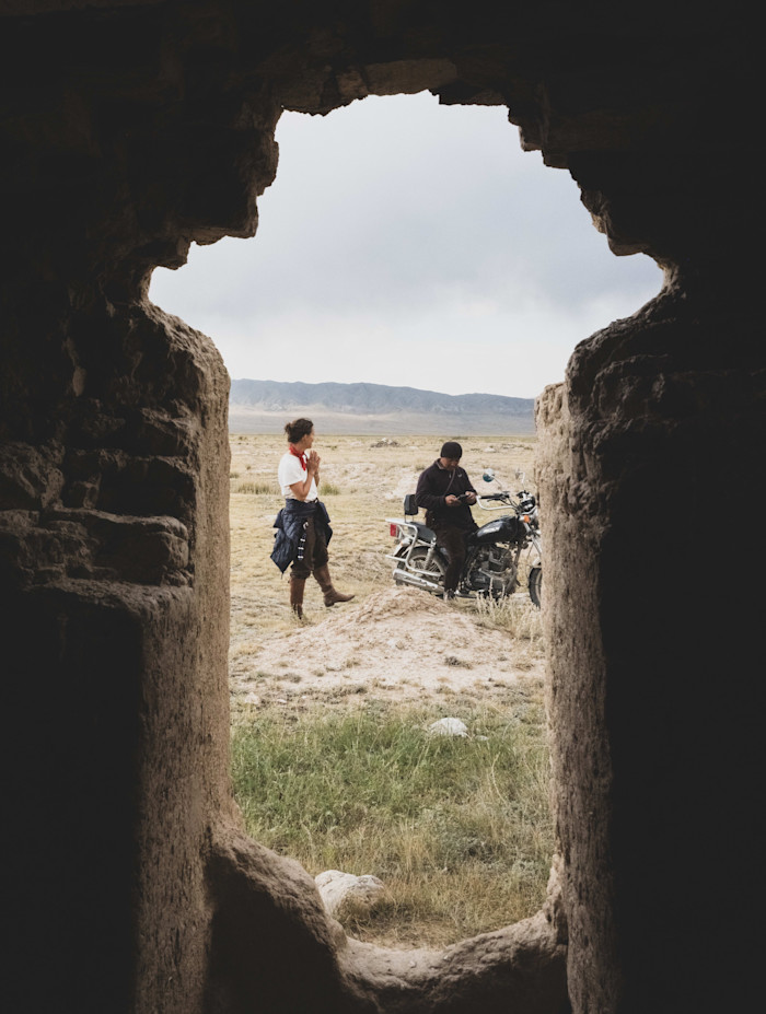 Two people are seen through a stone window; one stands and another sits on a motorbike, talking in a grassy, remote landscape.