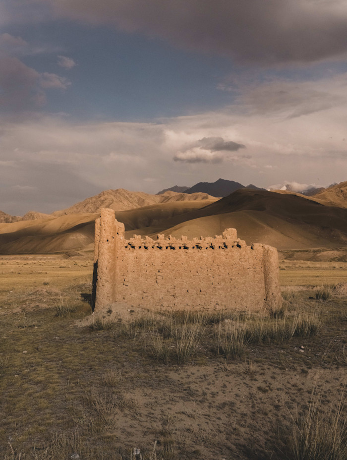 A crumbling mud-brick cemetery building stands alone on a grassy plain with mountains in the background.
