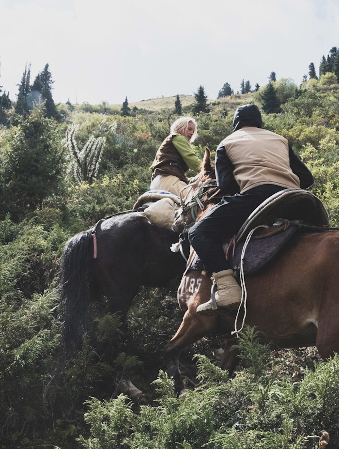 Alexandra Tolstoy rides a horse up a steep, grassy incline, assisted by a Kyrgyz riding guide on horseback ahead.