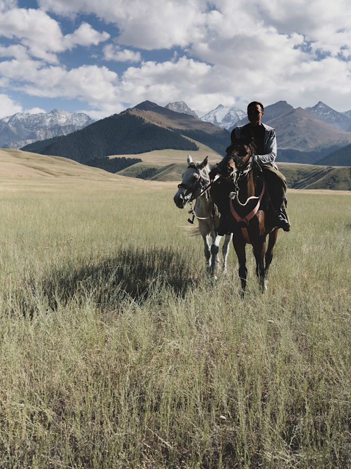 Adil rides a horse through a grassy plain with another horse beside him, snow-capped mountains in the background.