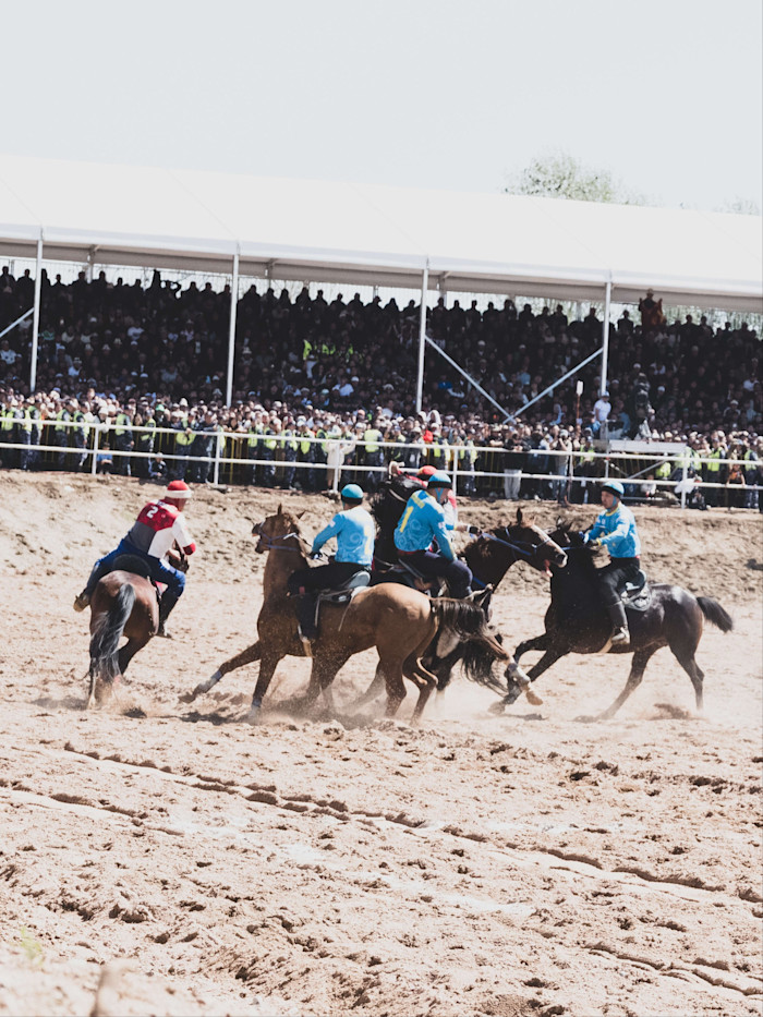 Players on horseback compete in a kok-boru match on a dusty field, with a large crowd watching from shaded stands.